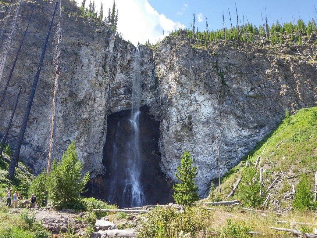 A real photograph of Fairy Falls in Yellowstone with a narrow waterfall dropping over a rock ledge into a forested basin, summer greenery and mist at the base