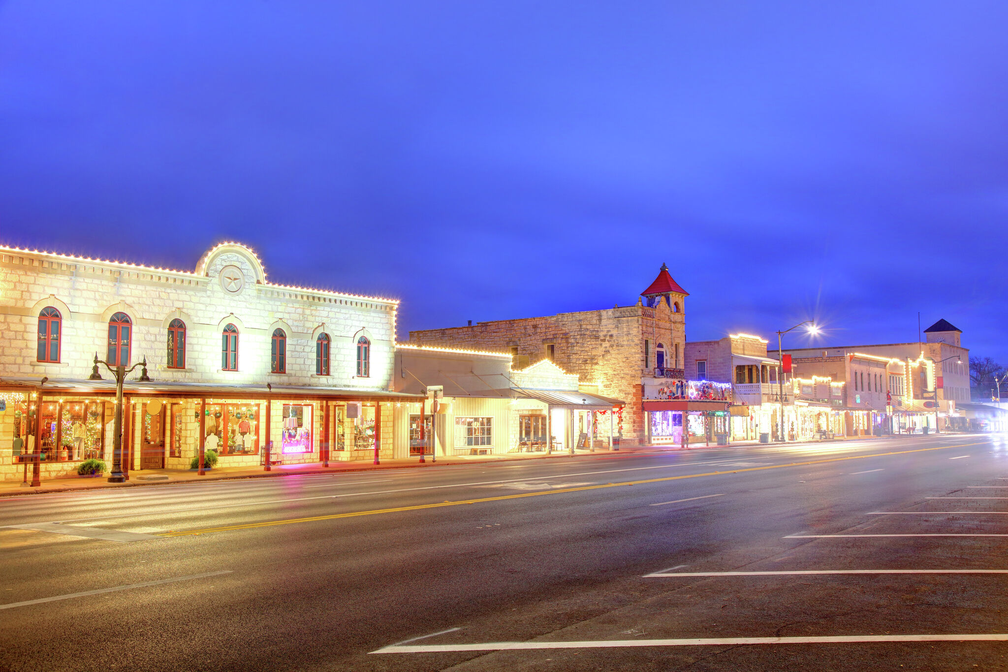 A real photograph of Fredericksburg, Texas on a warm evening, with historic storefronts along Main Street, people strolling on the sidewalks, and soft golden light reflecting off shop windows