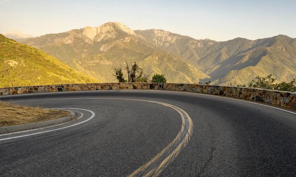 A real photograph of Generals Highway winding through dense conifer forest in Sequoia National Park with a car rounding a curve in late afternoon