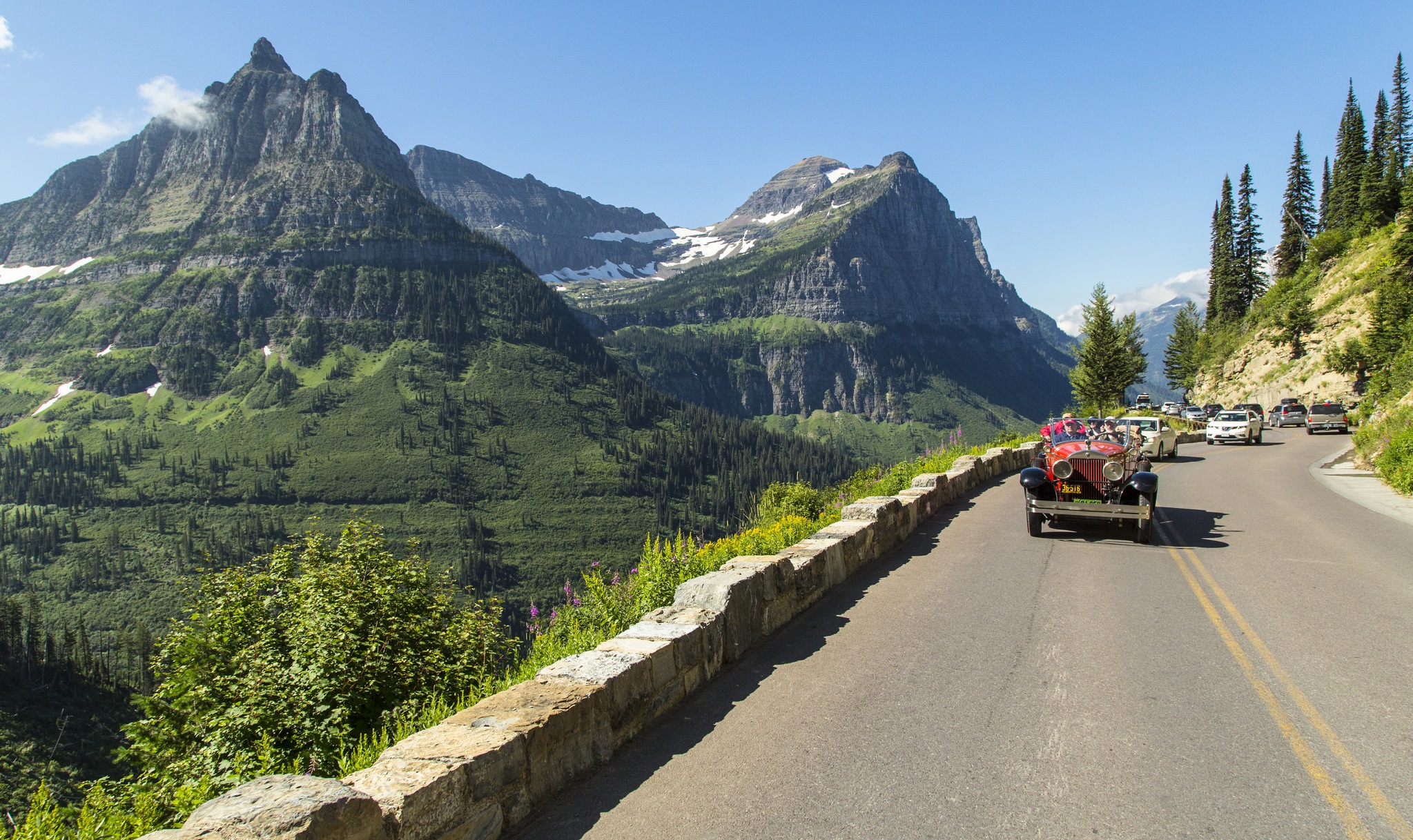 A real photograph of Going-to-the-Sun Road in Glacier National Park on a clear summer day, viewed from a roadside overlook with a stone guardrail, jagged mountain peaks and a snow-streaked ridge in the background, and a few cars winding along the narrow mountain road, natural colors, photorealistic