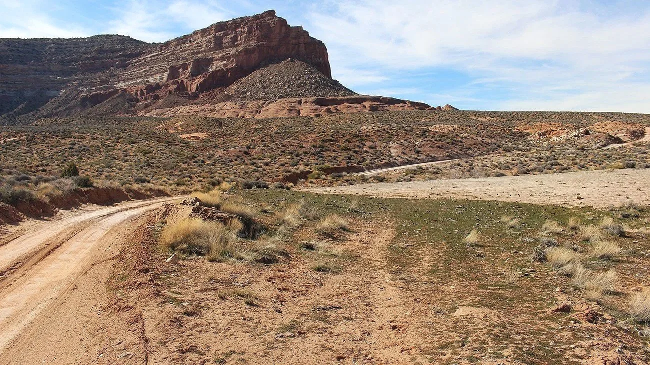 A real photograph of Hole-in-the-Rock Road in Grand Staircase–Escalante, showing a dusty washboard dirt road stretching toward slickrock hills under a clear sky