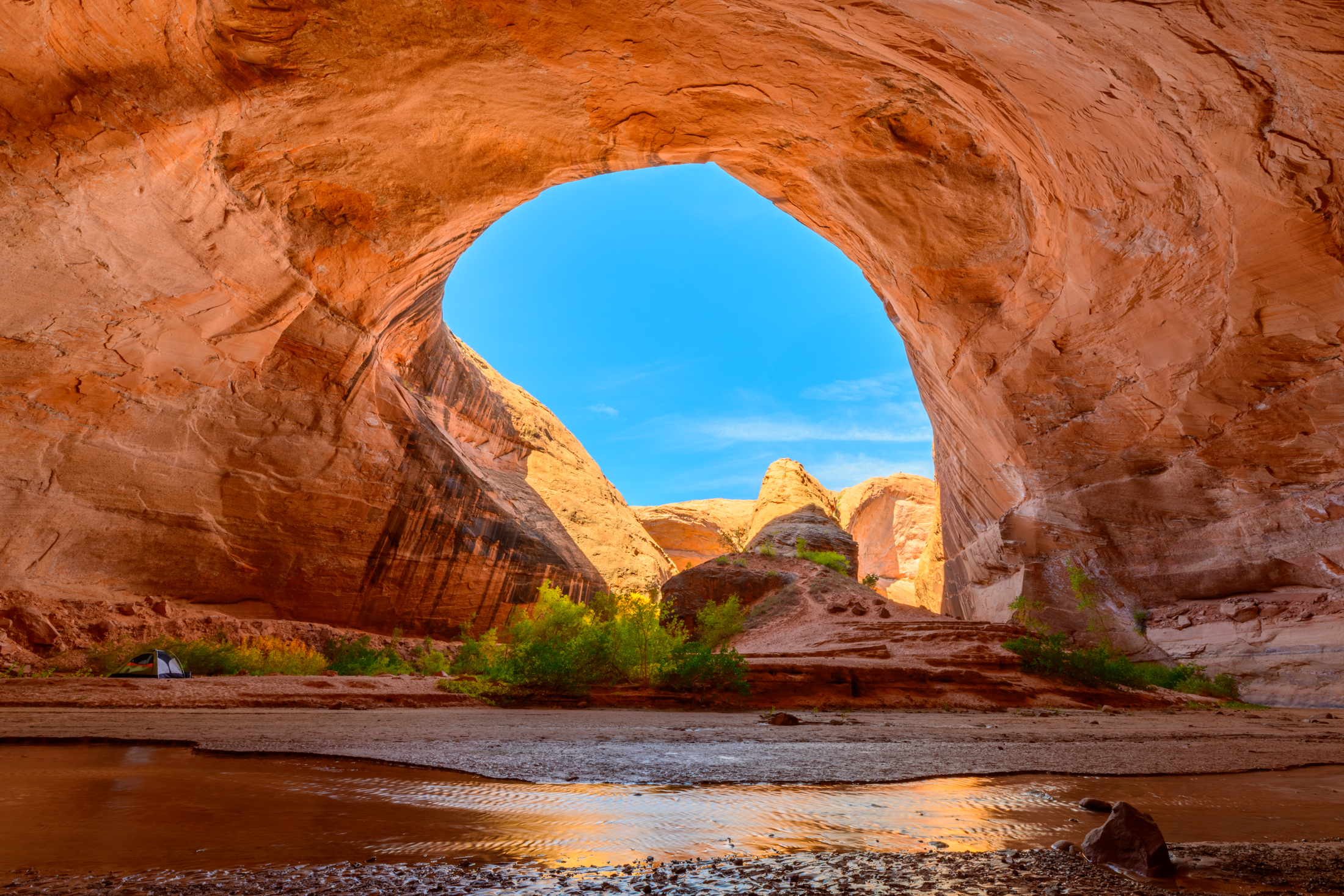 A real photograph of Jacob Hamblin Arch in Coyote Gulch, with a hiker standing near the stream beneath the massive sandstone arch, cottonwoods lining the canyon floor
