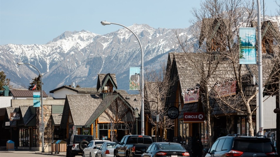 A real photograph of Jasper town streets with low wooden buildings, a few parked cars, and distant Rocky Mountain peaks under a clear blue sky, calm small-town atmosphere