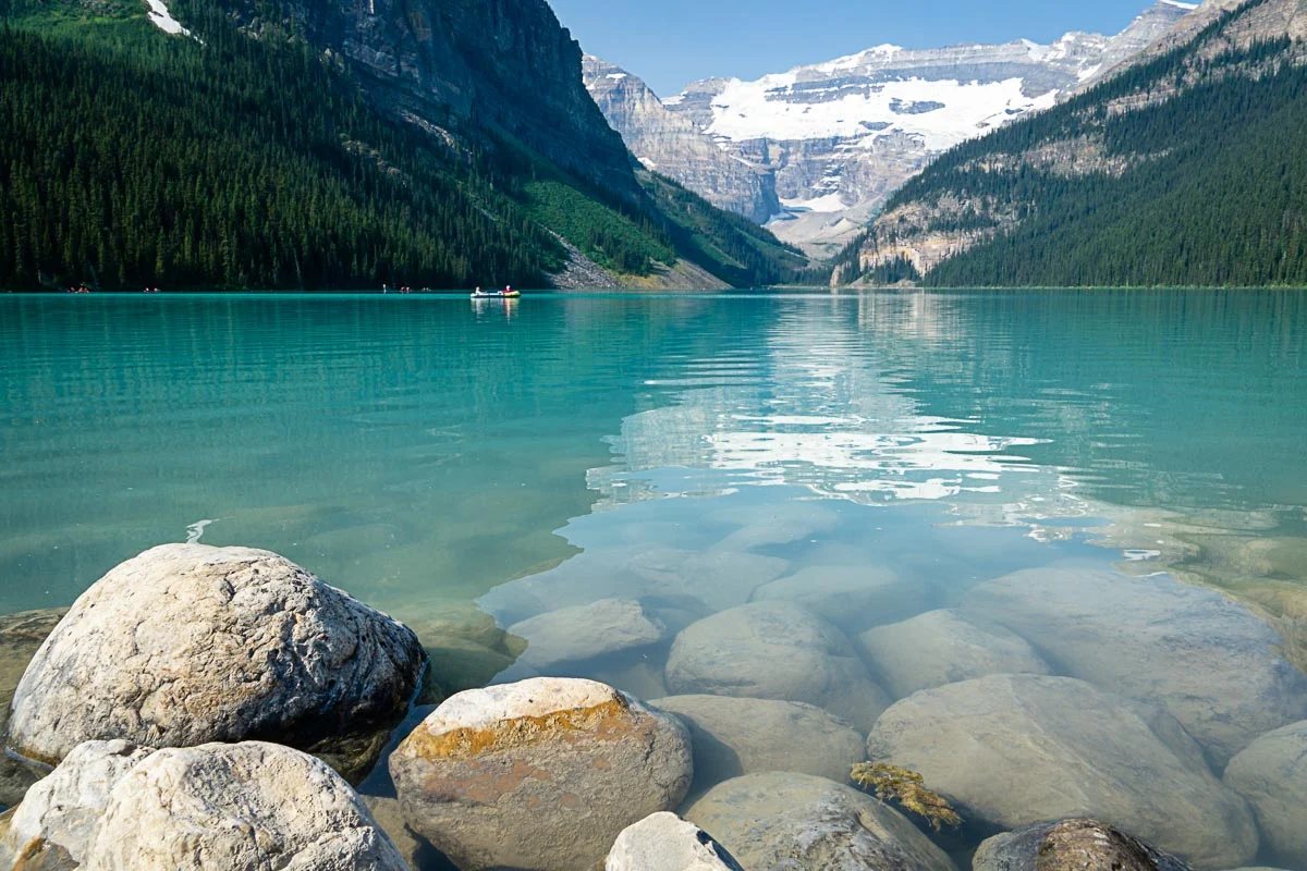 A real photograph of Lake Louise on a sunny summer day with turquoise water, canoes near the shore, and steep mountains rising behind the lake