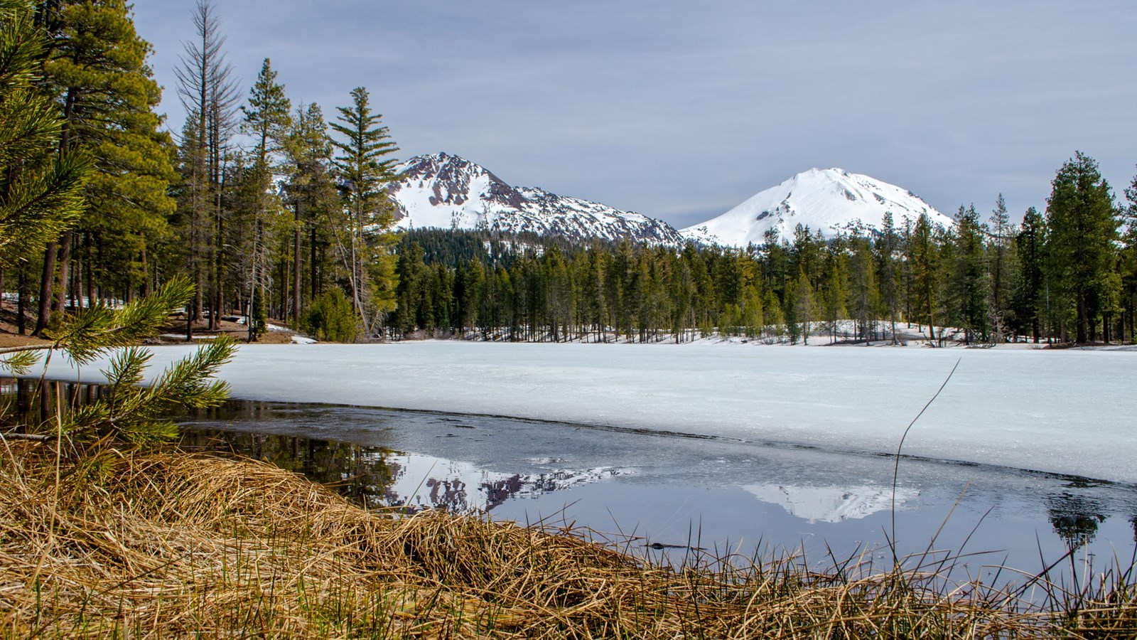 A real photograph of Lassen Peak reflected in the calm water of Manzanita Lake with pine trees framing the shoreline in Lassen Volcanic National Park