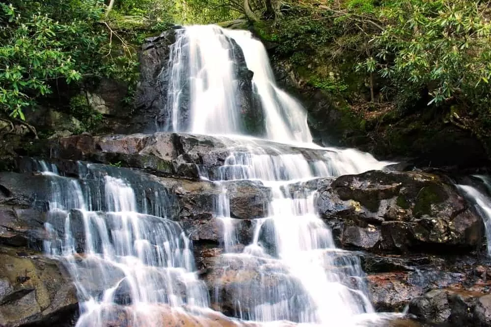 A real photograph of Laurel Falls in Great Smoky Mountains National Park, water cascading over a rocky ledge into a shallow pool with mossy boulders and green forest surrounding the scene