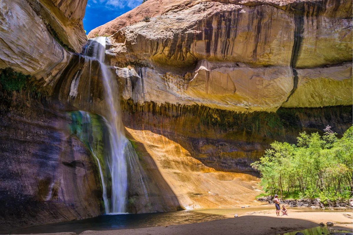 A real photograph of Lower Calf Creek Falls dropping into a green pool with hikers resting on sandy shoreline and towering red canyon walls, bright desert afternoon
