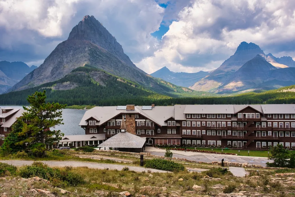 A real photograph of Many Glacier Hotel viewed from the shoreline of Swiftcurrent Lake in the morning light, with calm water reflecting mountains
