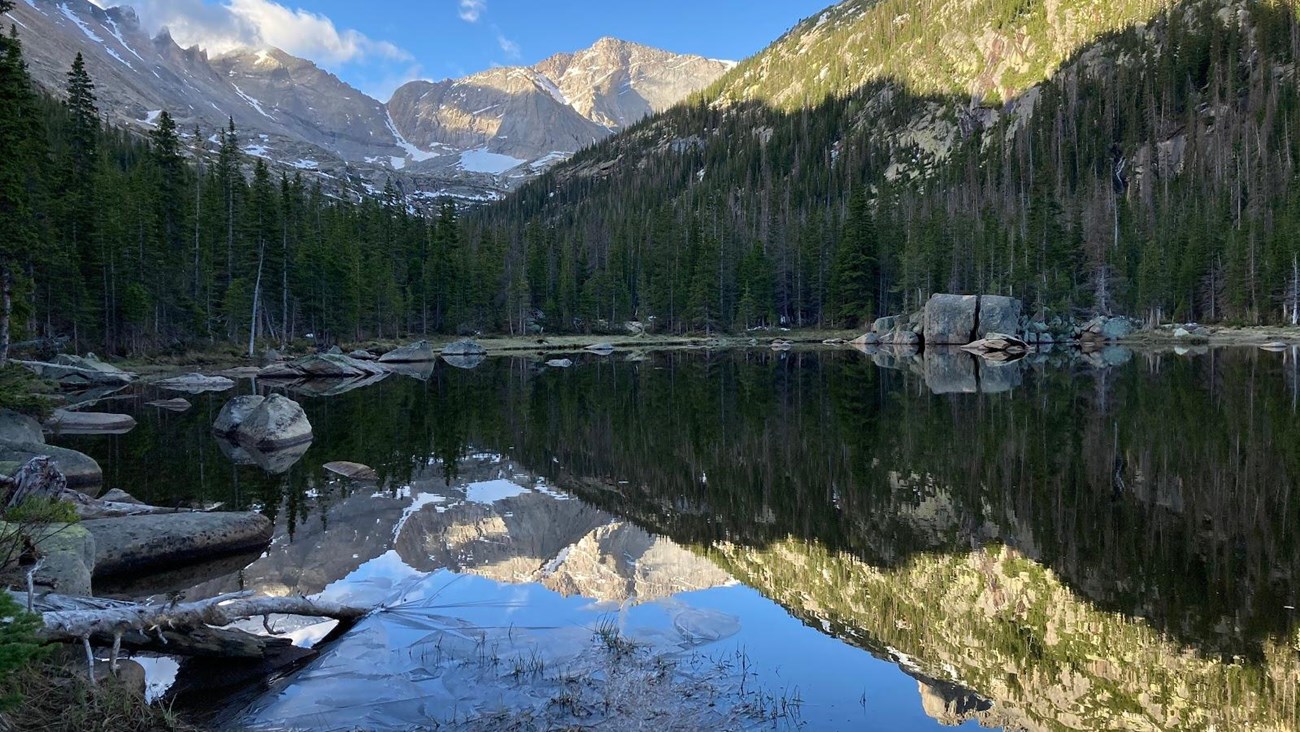 A real photograph of Mills Lake with hikers resting on rocks near the shoreline, evergreen trees framing the water, and Longs Peak looming in the distance