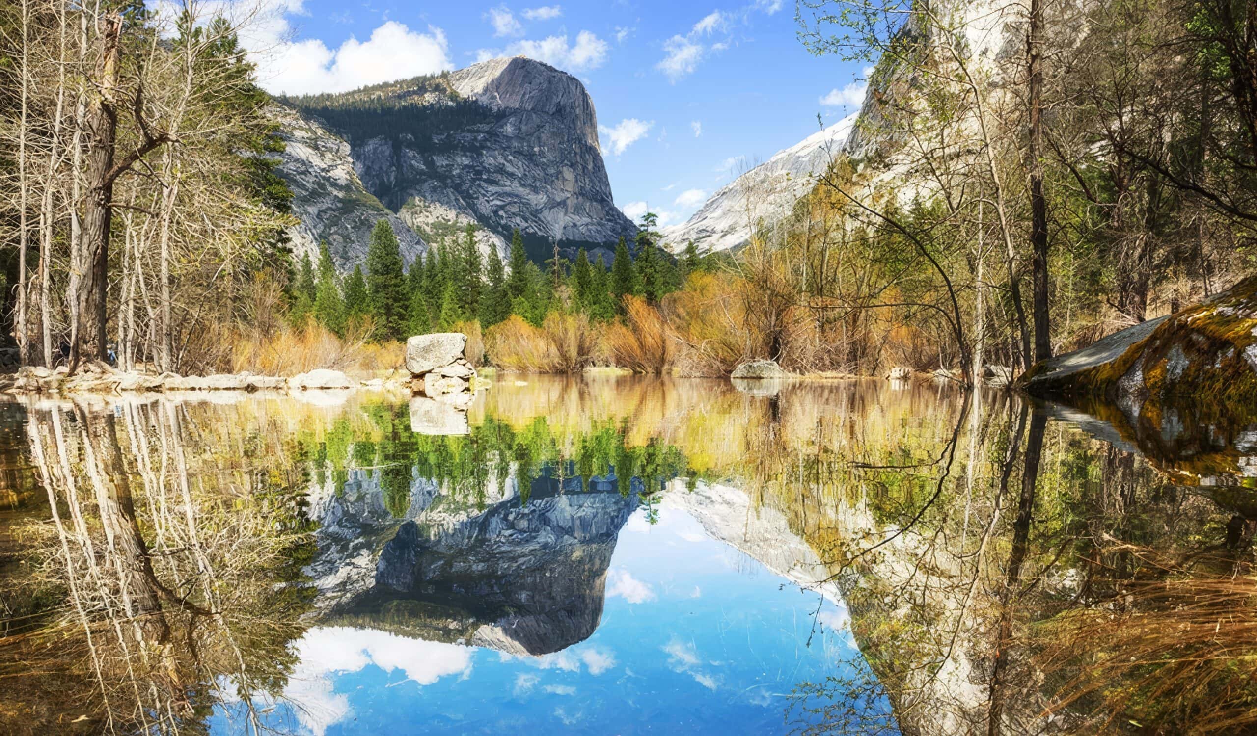 A real photograph of Mirror Lake in Yosemite National Park in spring, with calm water reflecting the face of Half Dome and leafy trees along the shoreline