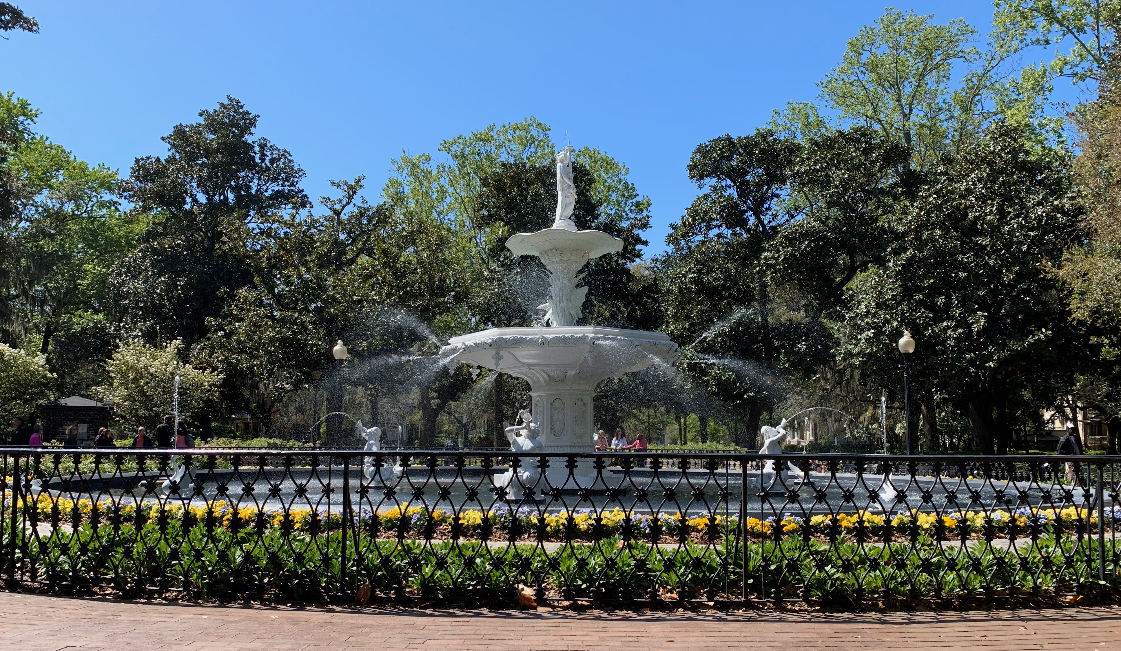 A real photograph of Monterey Square in Savannah with a central fountain, brick paths, and live oak trees covered in Spanish moss on a bright morning