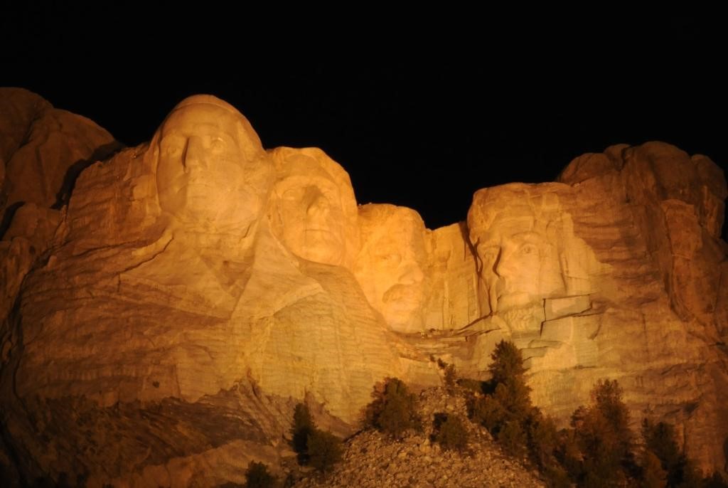A real photograph of Mount Rushmore at golden hour with warm light on the carved presidential faces and pine-covered Black Hills in the foreground
