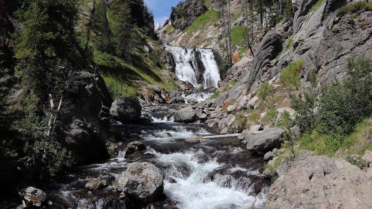 A real photograph of Mystic Falls in Yellowstone with a waterfall cascading through a forested setting, with hikers on a nearby trail