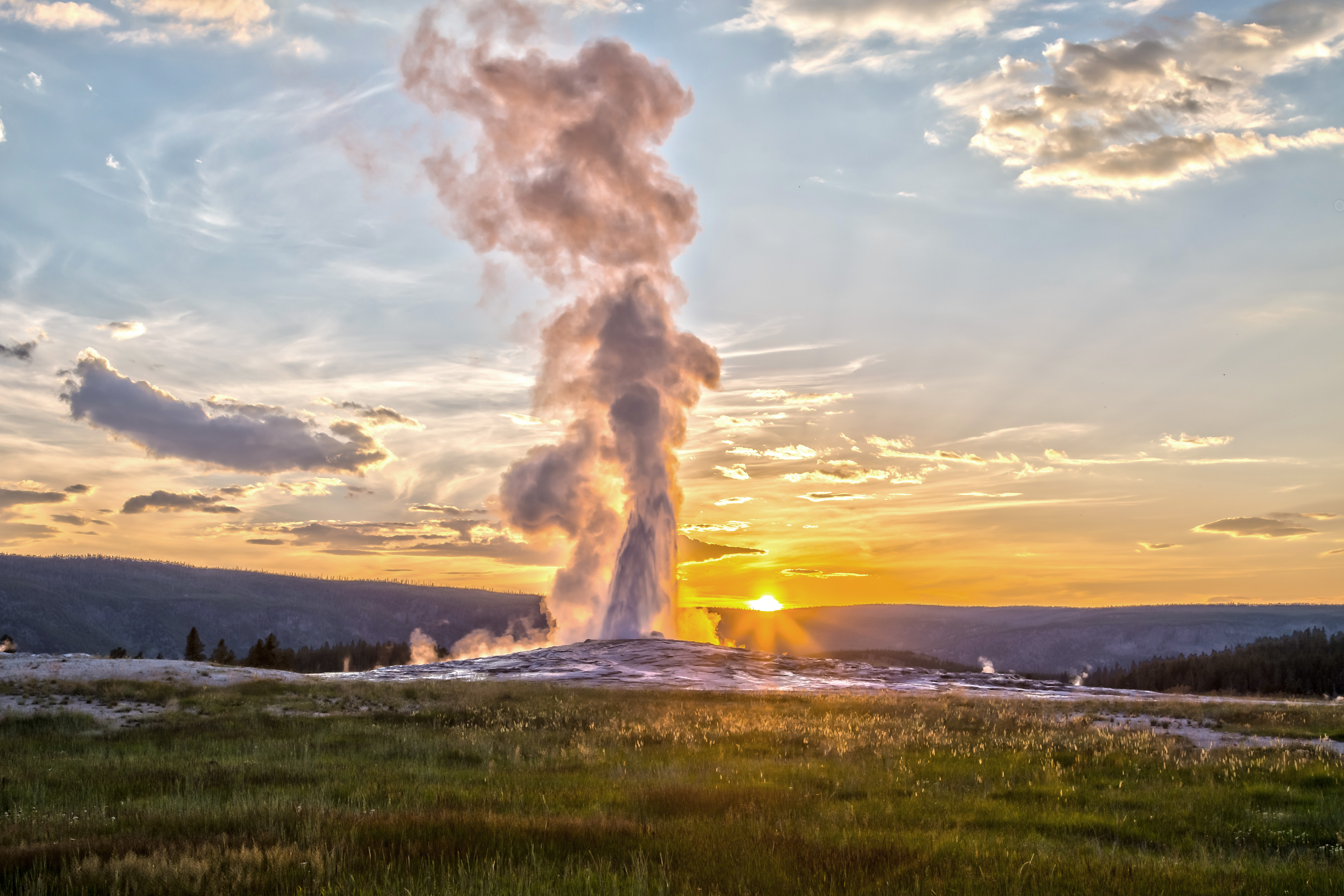 A real photograph of Old Faithful erupting in Yellowstone with a tall column of water and steam against a blue sky, visitors watching from a distance