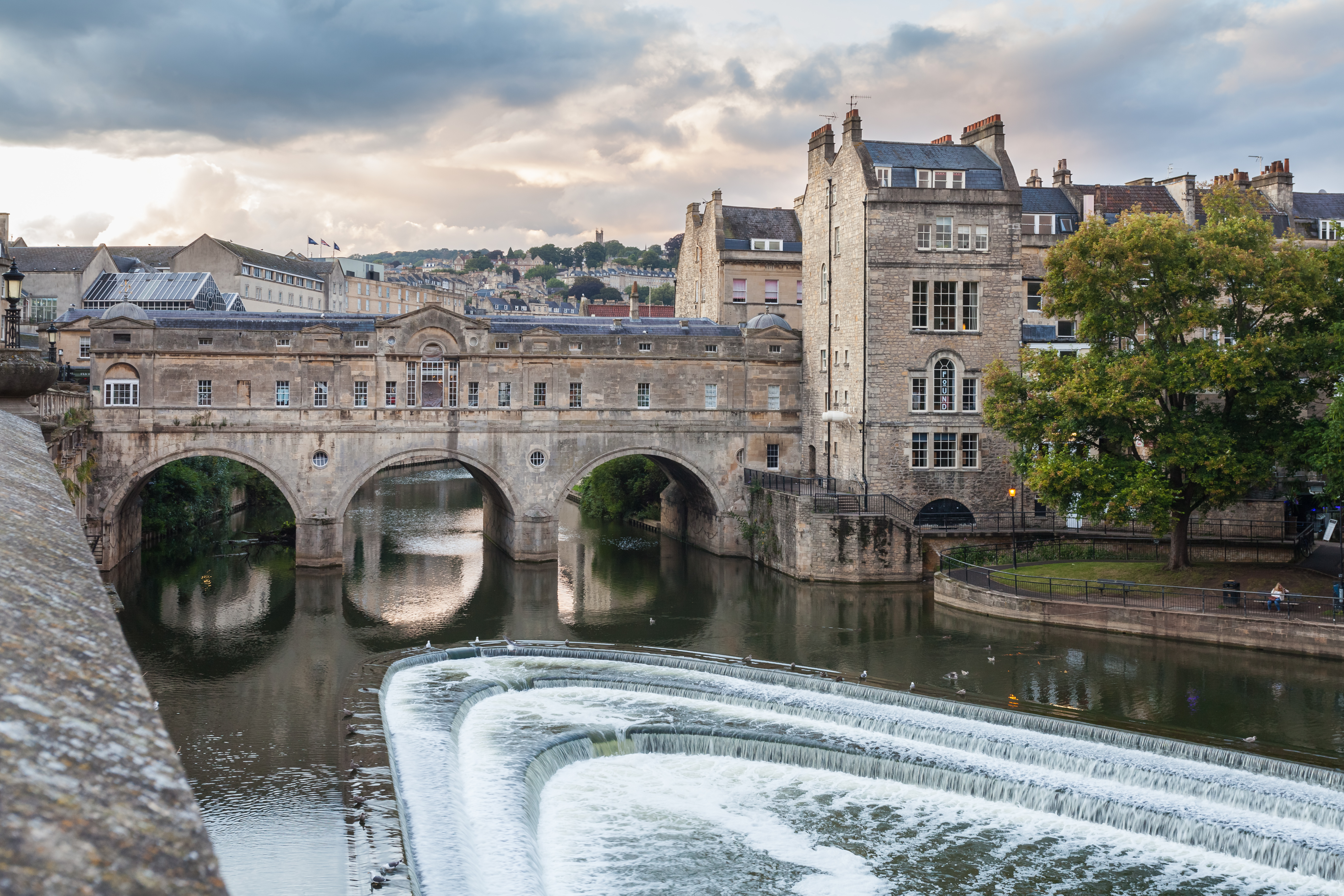 A real photograph of Pulteney Bridge in Bath viewed from the riverbank, showing the arched bridge with buildings on top and water flowing over the weir, late morning light, travel photography