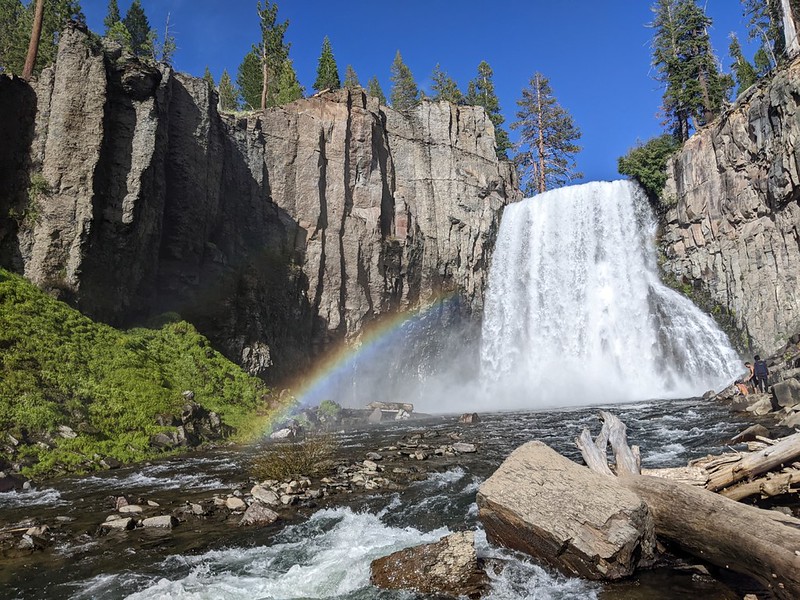 A real photograph of Rainbow Falls near Devils Postpile, with white water cascading over a rocky ledge into a pool and mist rising in bright daylight