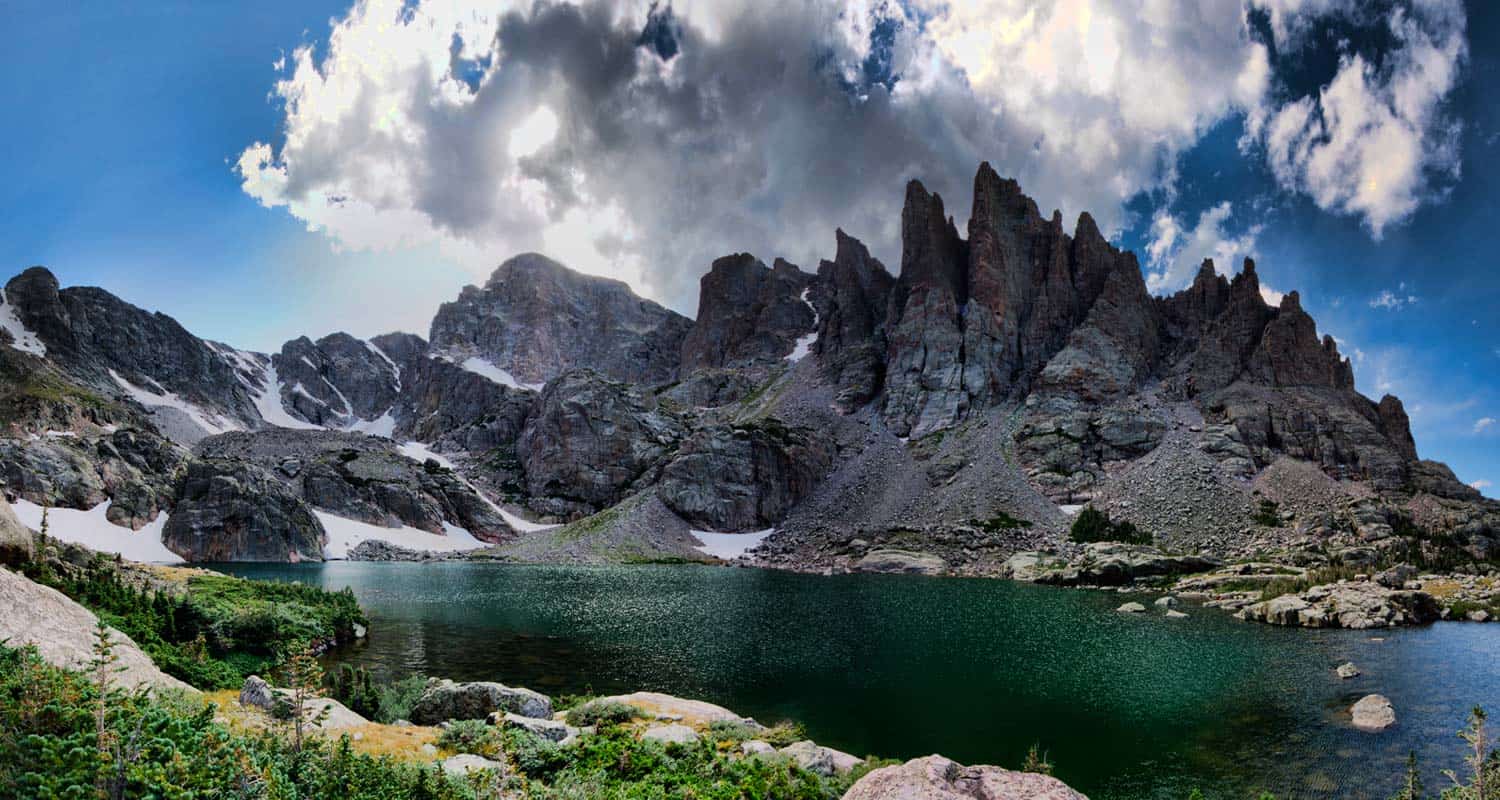 A real photograph of Sky Pond in Rocky Mountain National Park with clear water, jagged mountain spires, and hikers standing on boulders near the shore
