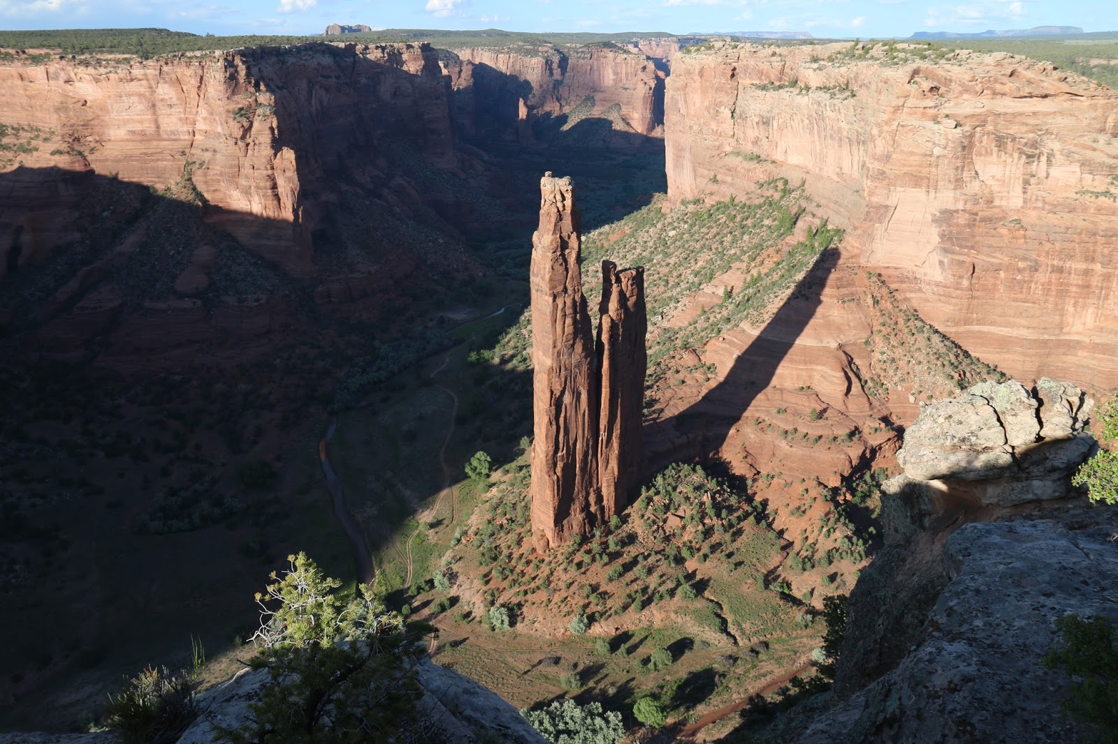 A real photograph of Spider Rock rising from the canyon floor as seen from a South Rim overlook at Canyon de Chelly, with layered red sandstone walls and a wide desert sky