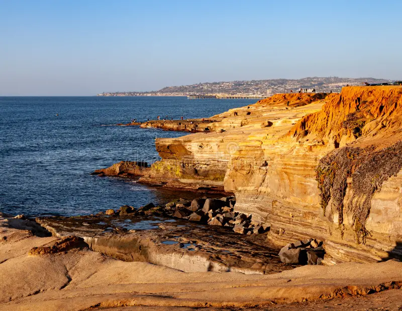 A real photograph of Sunset Cliffs in San Diego at golden hour, with waves breaking against rugged coastal rock and a few people watching from the bluff