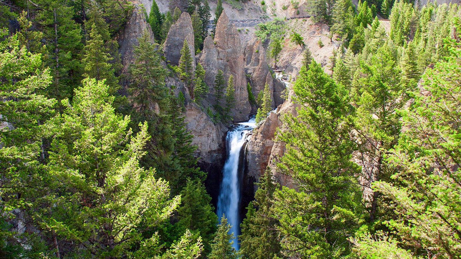 A real photograph of Tower Fall in Yellowstone dropping between rugged rock spires, with a green forested canyon below and bright daylight