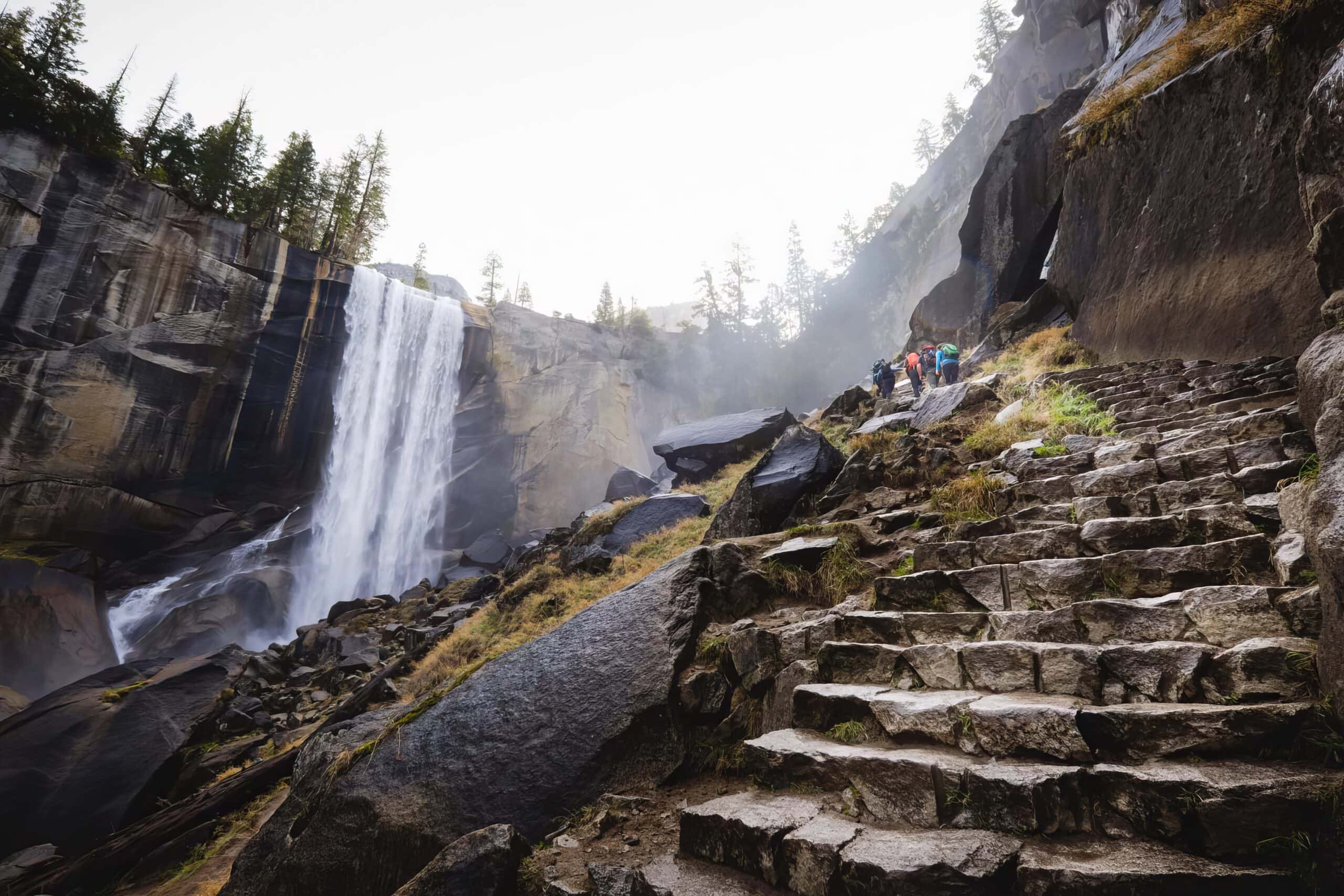 A real photograph of Vernal Fall in Yosemite pouring over a granite cliff, with mist rising and hikers visible on the stone steps of the Mist Trail, bright daylight