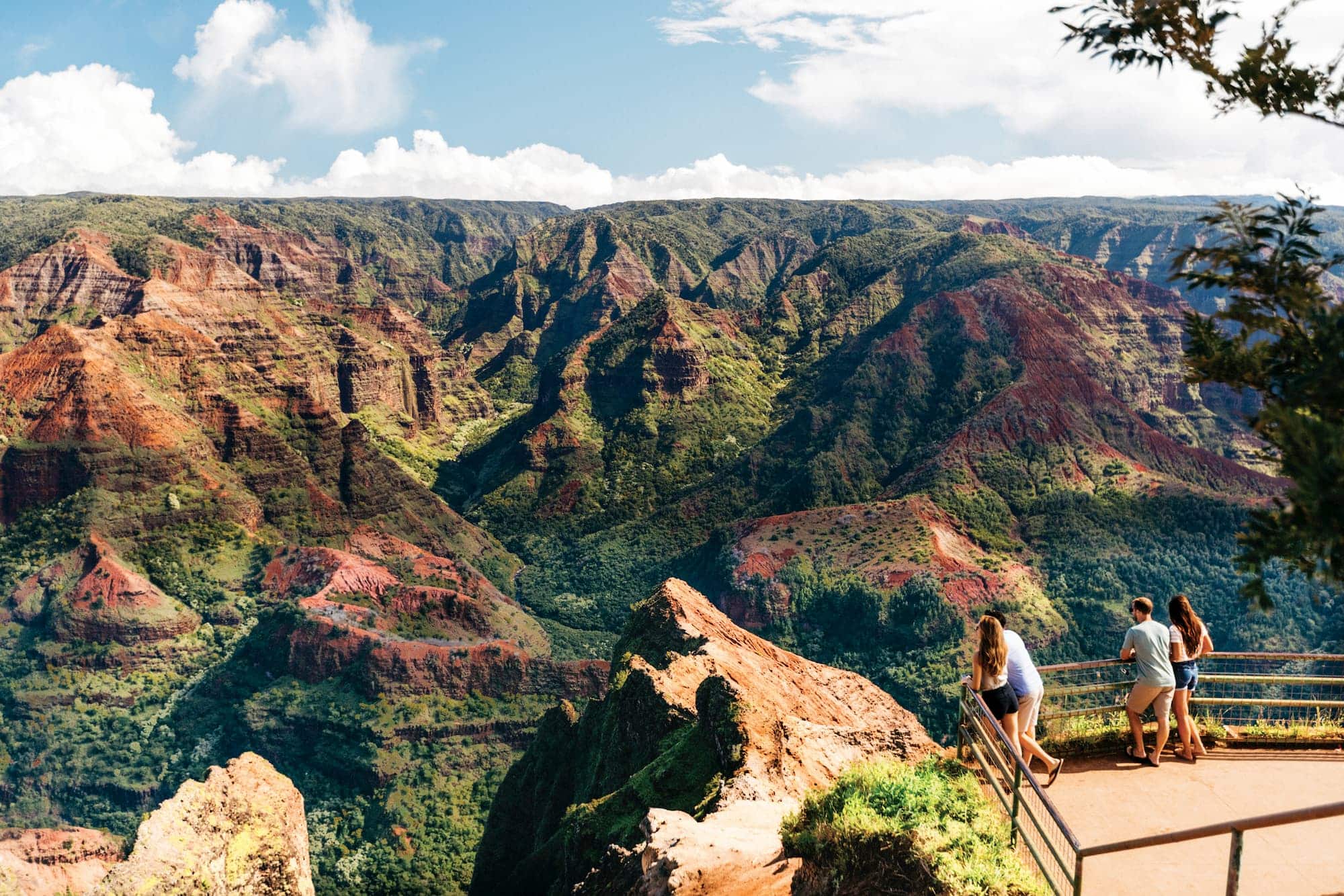 A real photograph of Waimea Canyon in Kauai seen from a roadside lookout, with red and green canyon walls and layered ridgelines under broken afternoon clouds