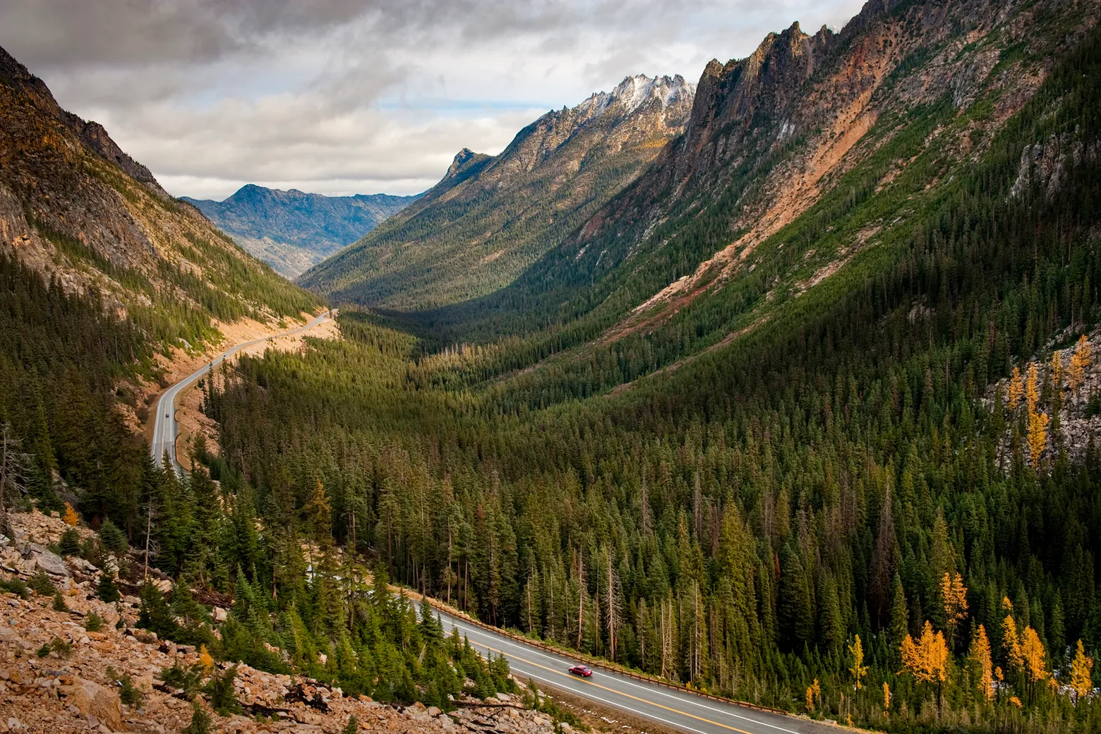 A real photograph of Washington State Route 20 winding through steep, forested North Cascades mountains on a clear summer day, with a wide scenic overlook and distant jagged peaks
