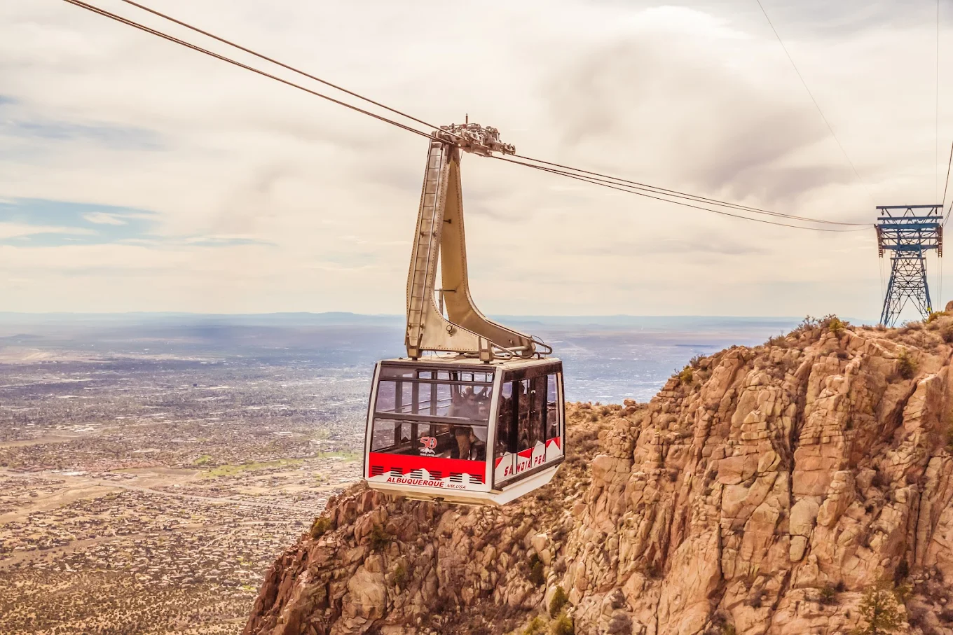 A real photograph of a Sandia Peak Tramway cabin suspended over a rugged canyon with the city visible in the distance, clear sky
