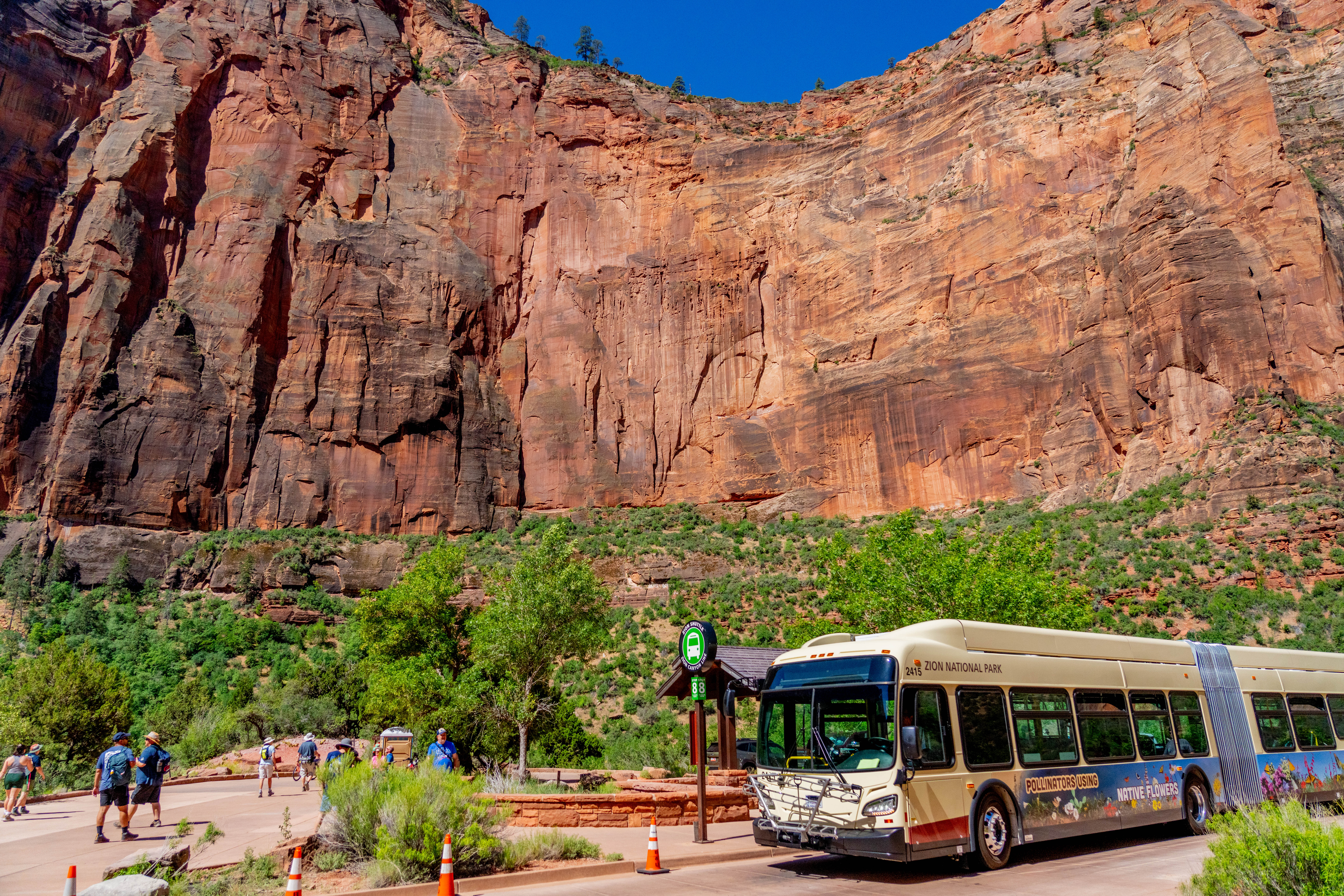 A real photograph of a Zion Canyon shuttle bus stopped near the Temple of Sinawava area with visitors stepping off near trail signage and canyon cliffs