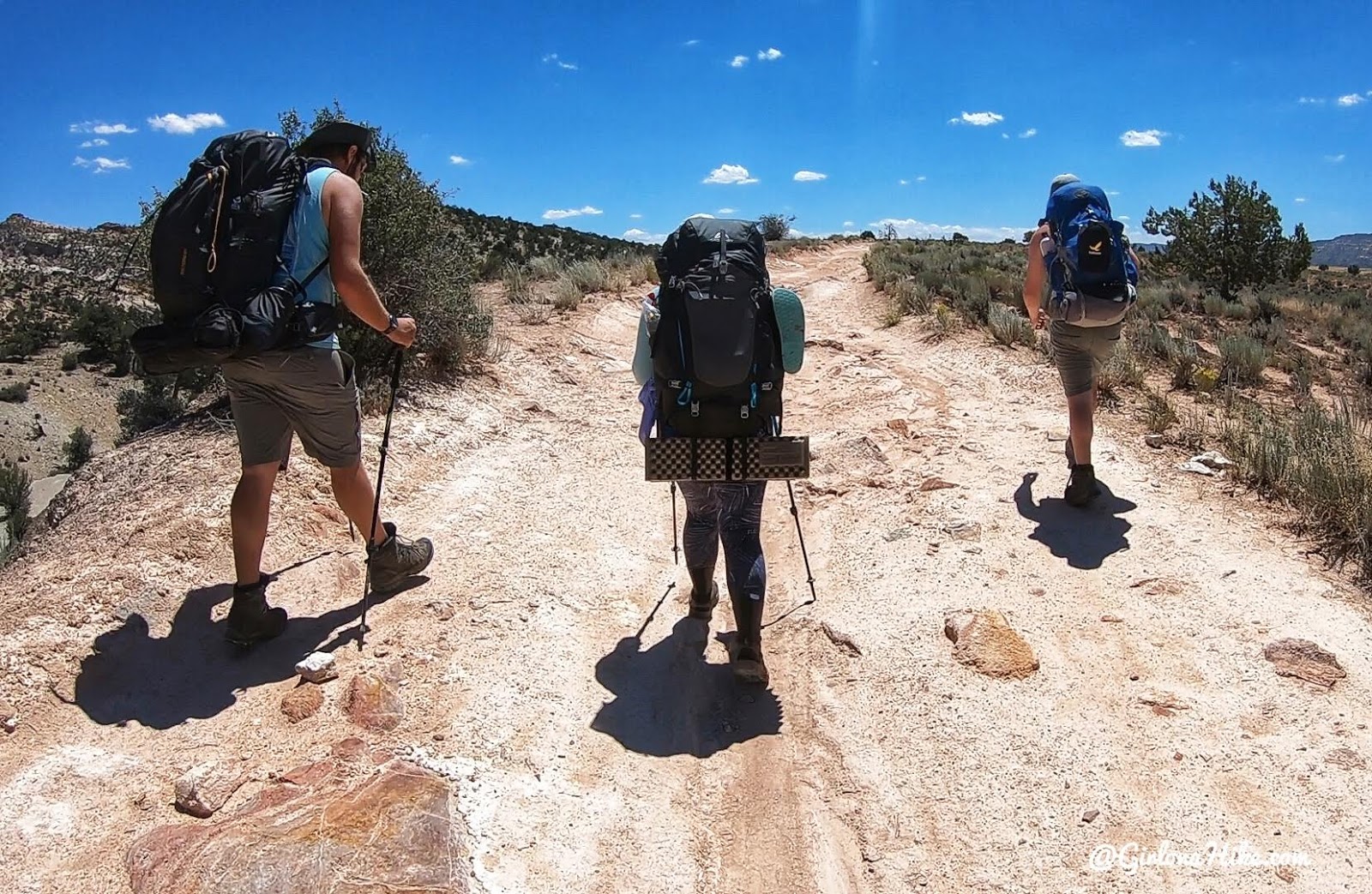 A real photograph of a backpack, trekking poles, and water bottles resting on sunlit slickrock near the edge of a desert canyon