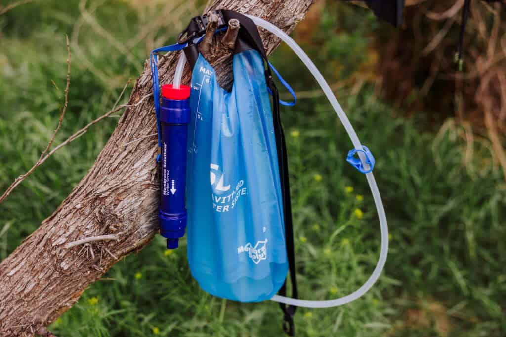 A real photograph of a backpacker crouched beside a clear stream near Havasu Creek, using a handheld water filter to fill a bottle, with canyon walls and cottonwood shade behind