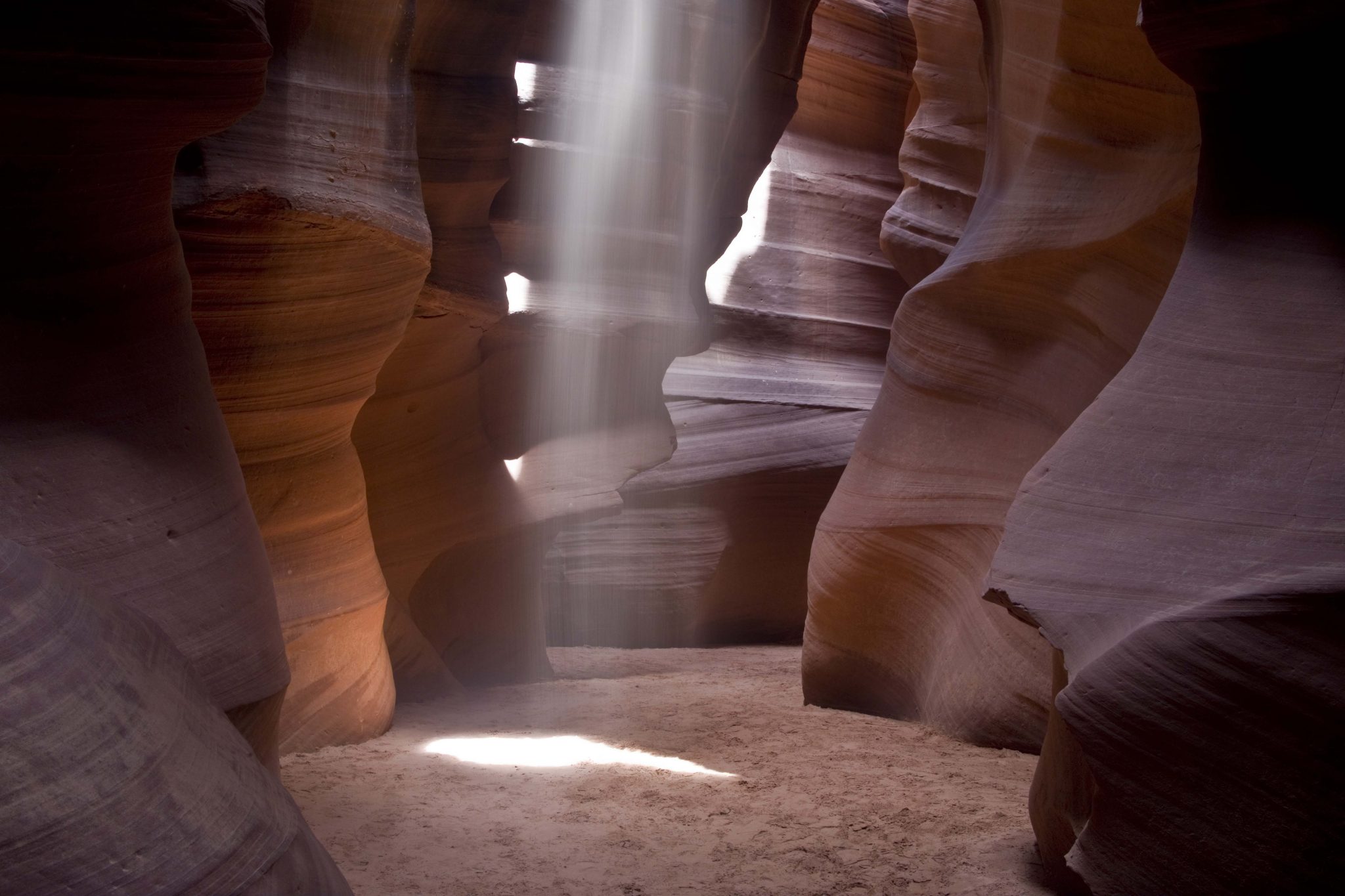 A real photograph of a bright sunbeam streaming into Upper Antelope Canyon, illuminating swirling orange sandstone walls and dusty air in Page, Arizona