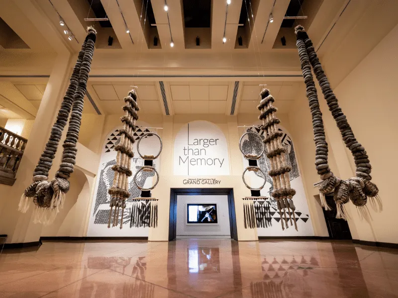 A real photograph of a calm gallery interior at the Heard Museum in Phoenix, with framed Indigenous artwork on neutral walls and soft museum lighting