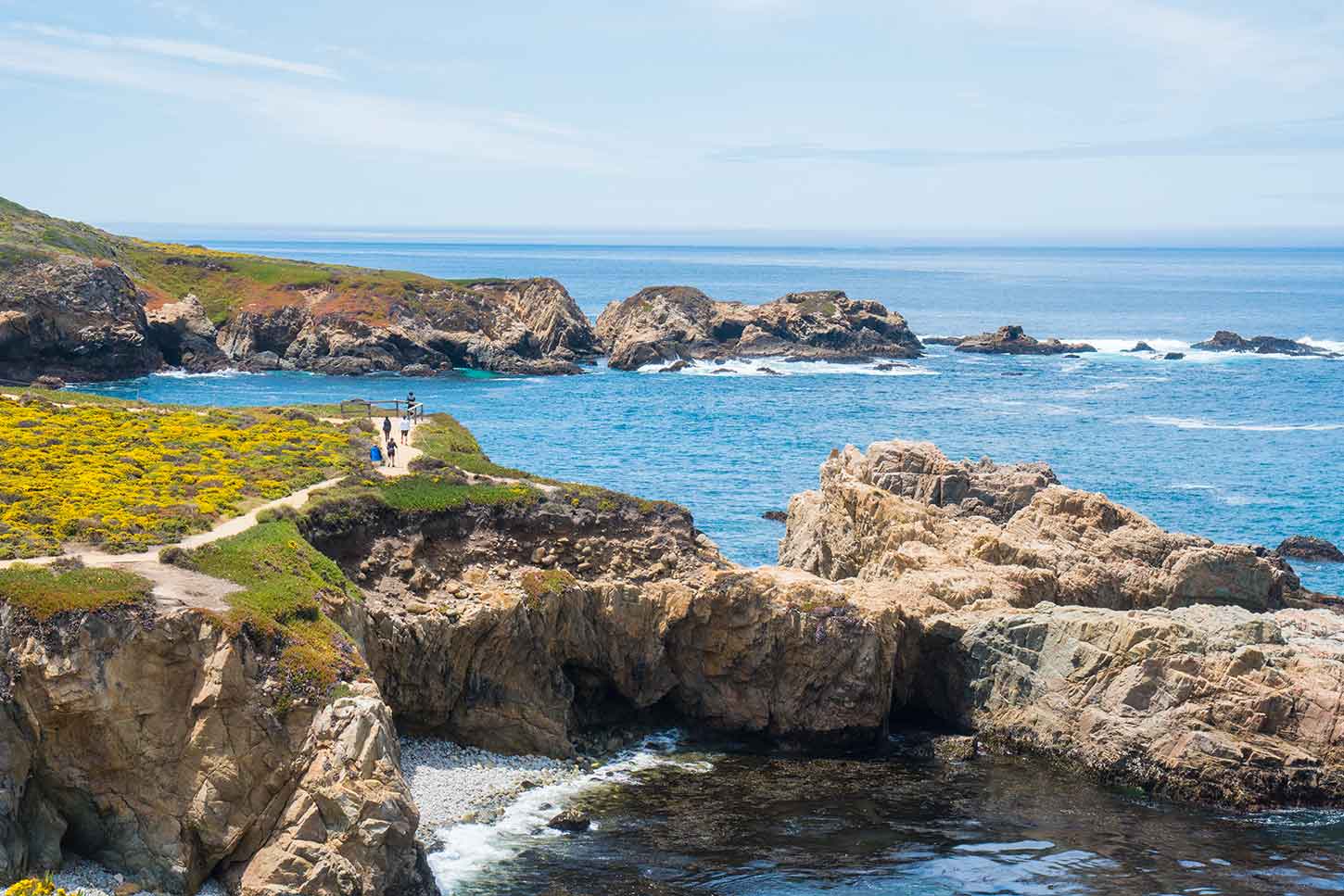 A real photograph of a coastal bluff trail at Garrapata State Park with golden grass, rugged cliffs, and white waves below under a blue sky