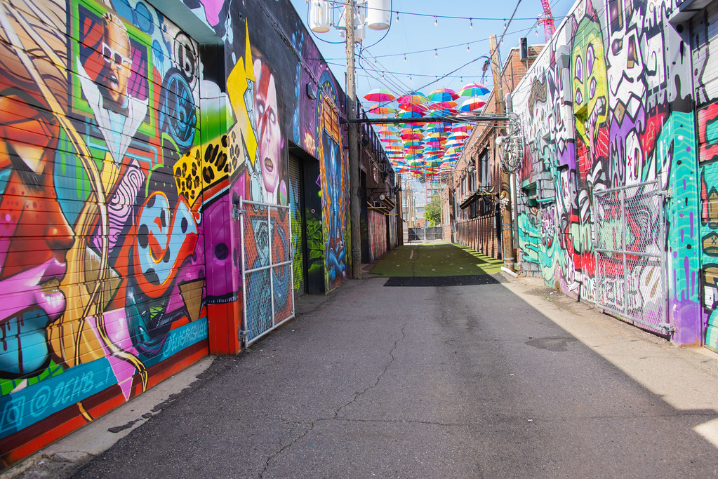 A real photograph of a colorful street art mural in an alley in Denver's RiNo Art District, with brick walls and a few pedestrians walking past