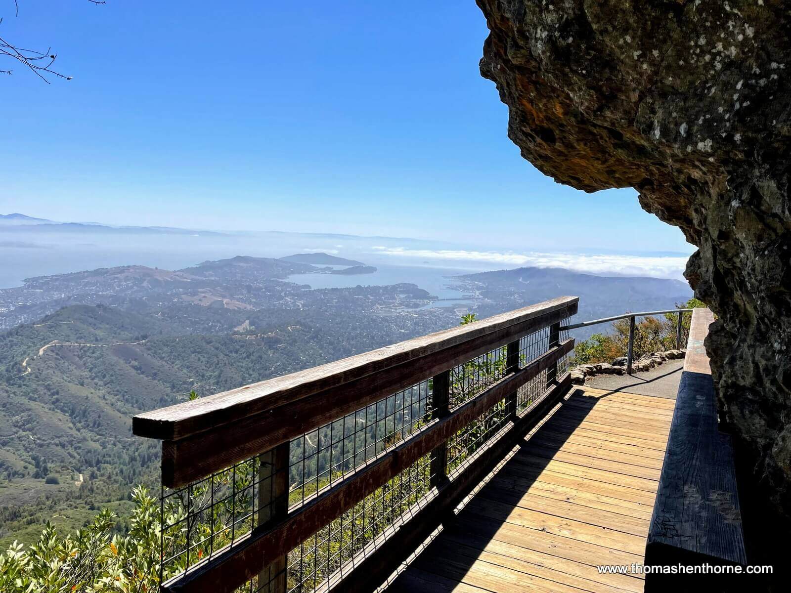 A real photograph of a dirt hiking trail on a Marin County ridgeline with coastal scrub, rolling hills, and a view toward the ocean under shifting fog