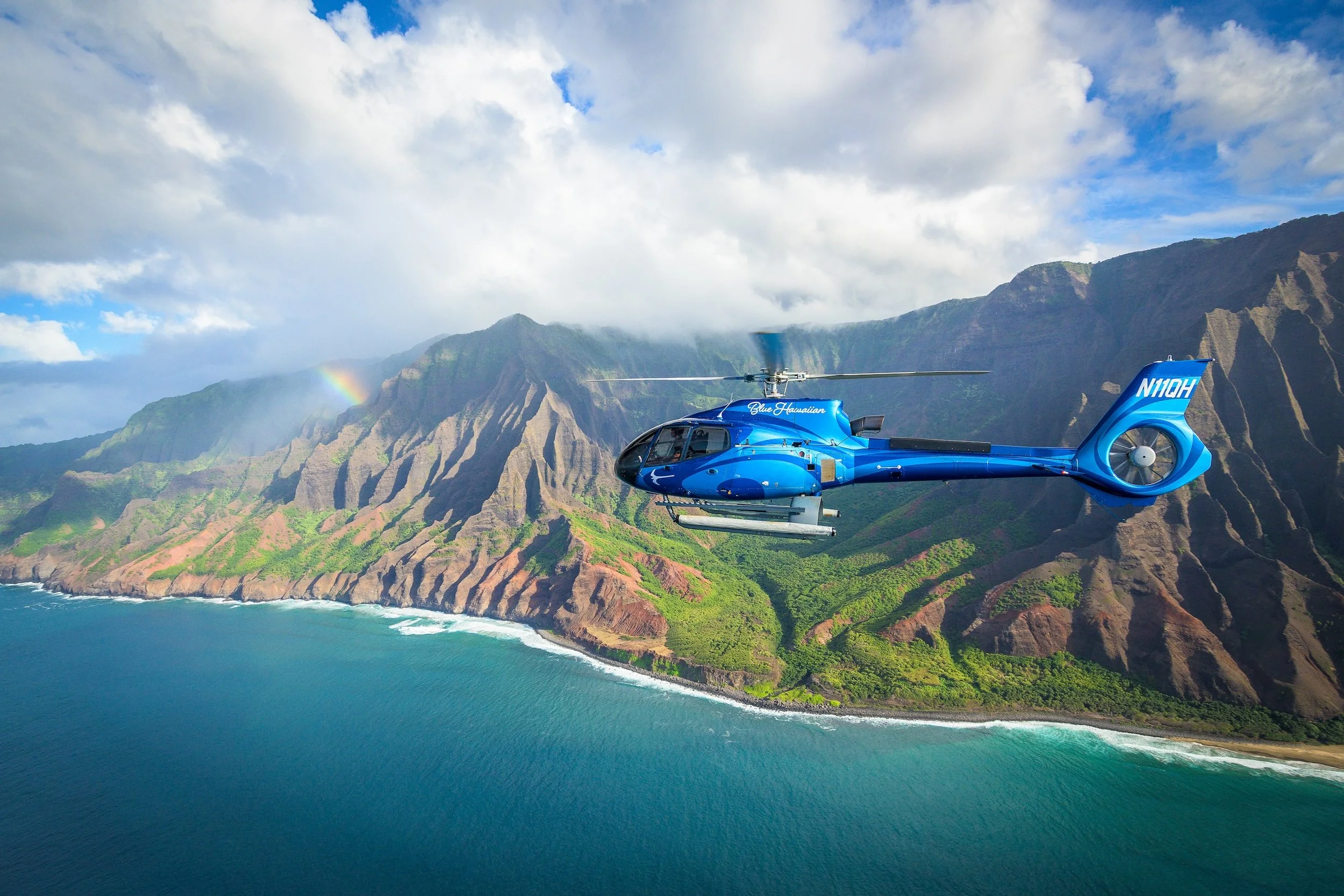 A real photograph of a helicopter flying above the Na Pali Coast on Kauai, with sharp green ridges and deep valleys visible below