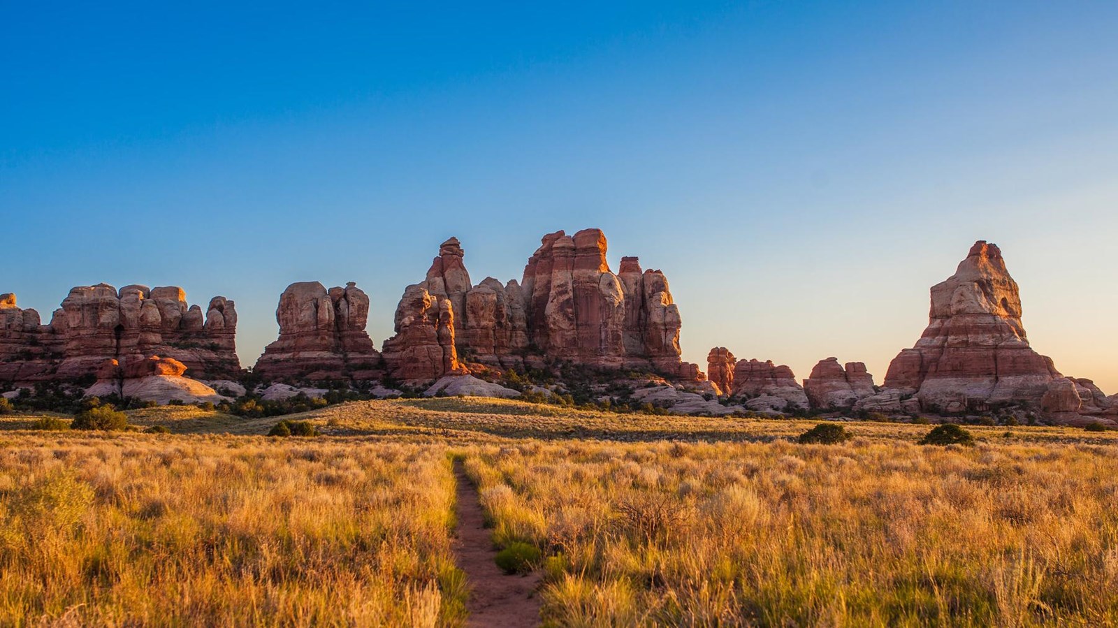 A real photograph of a hiker on the trail in Canyonlands Needles District near Chesler Park, surrounded by tall red-and-white banded sandstone spires under a clear sky