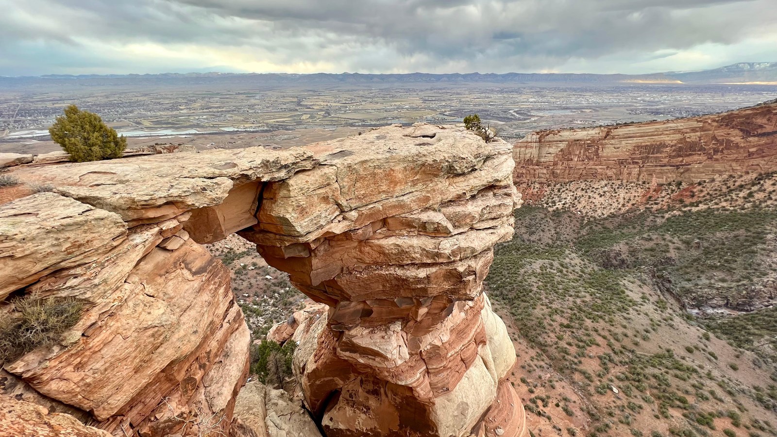 A real photograph of a hiker standing near a natural rock window on the Window Rock Trail in Colorado National Monument, with red sandstone framing the view