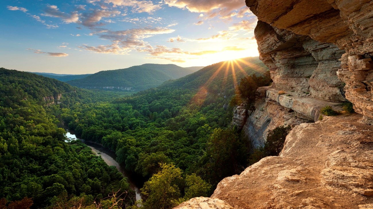 A real photograph of a hiker standing near the Big Bluff overlook above the Buffalo National River, with sweeping forested hills and a winding river far below