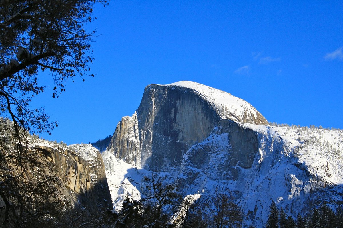 A real photograph of a hiker standing near the edge of the Half Dome summit in Yosemite National Park, looking out over the Valley on a clear afternoon