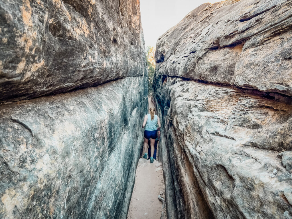 A real photograph of a hiker stepping through a narrow sandstone passage on the Joint Trail in Canyonlands Needles District, with tall rock walls close on both sides and soft reflected light