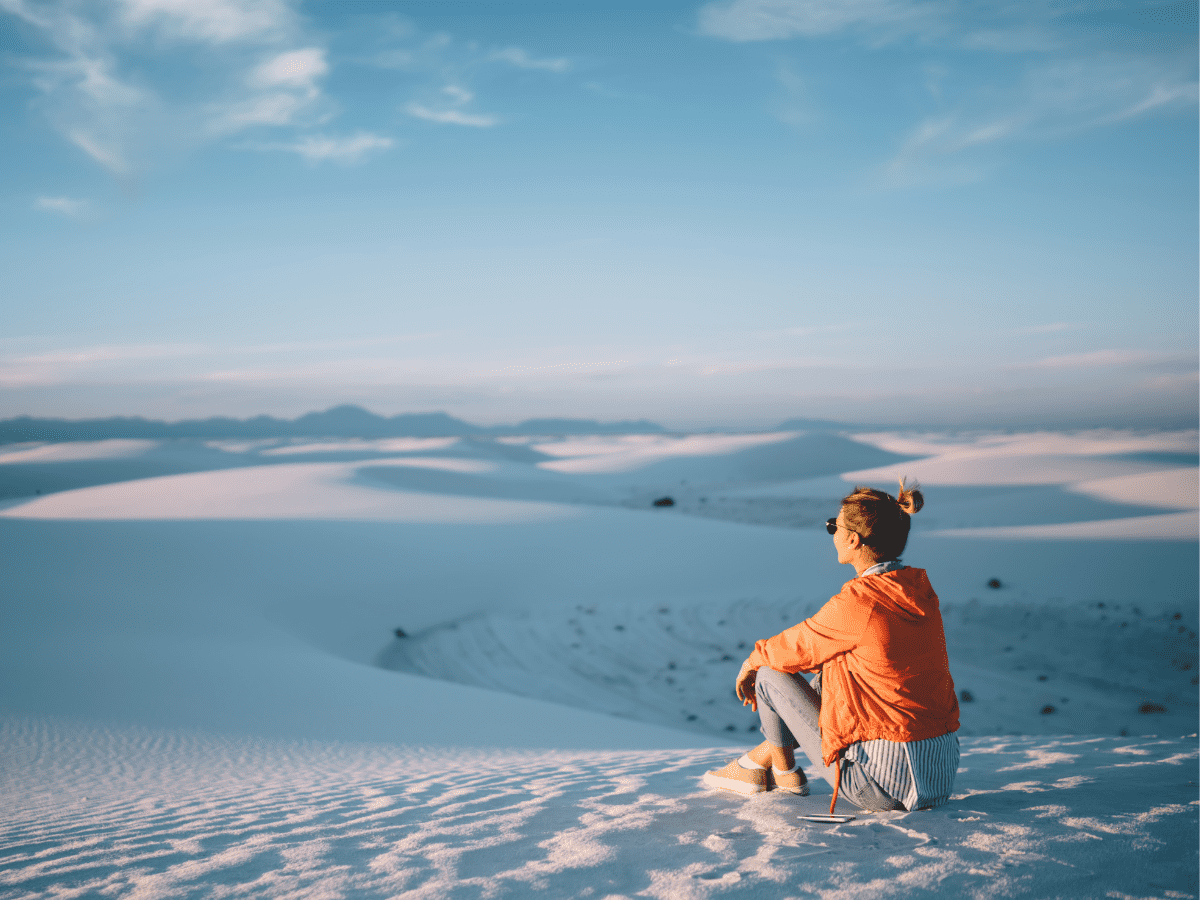 A real photograph of a hiker wearing a wide-brim hat and sunglasses walking across bright white gypsum dunes at White Sands National Park