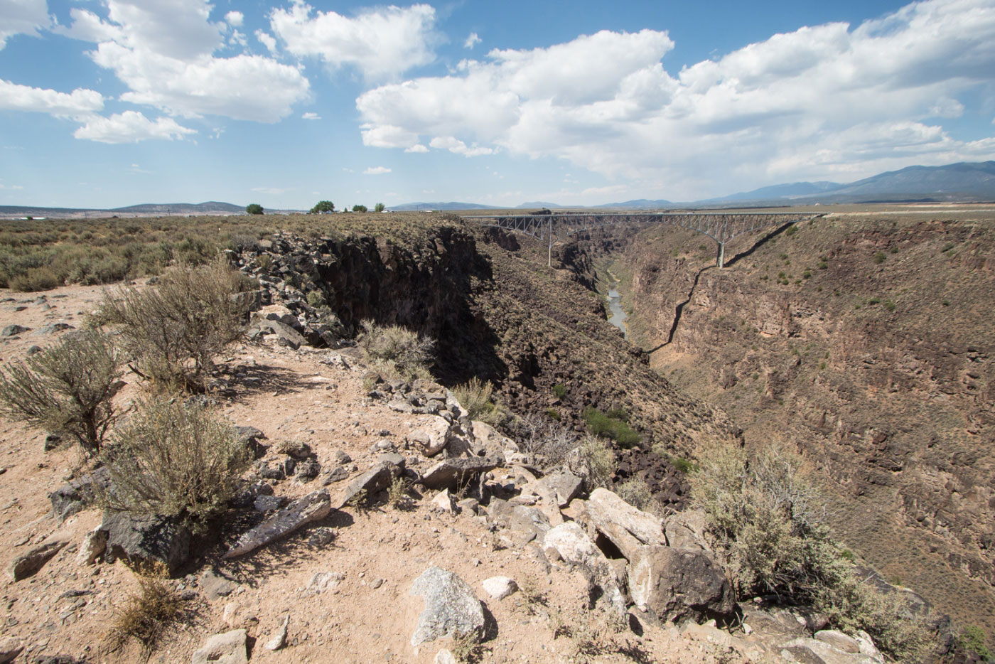 A real photograph of a narrow dirt trail along the high desert rim near Taos, New Mexico, with sagebrush, volcanic rock, and distant canyon views