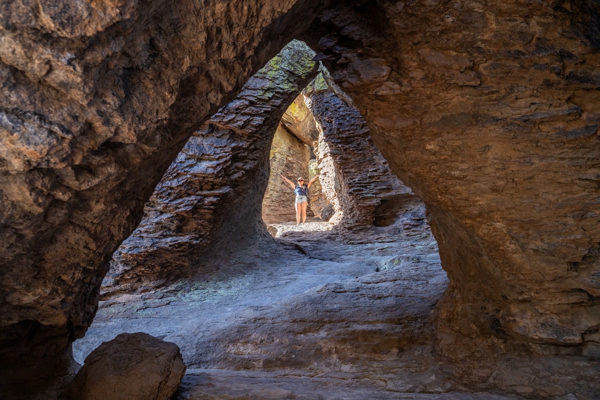 A real photograph of a narrow hiking trail winding between tall hoodoo rock walls at Chiricahua National Monument, with a hiker in the distance for scale