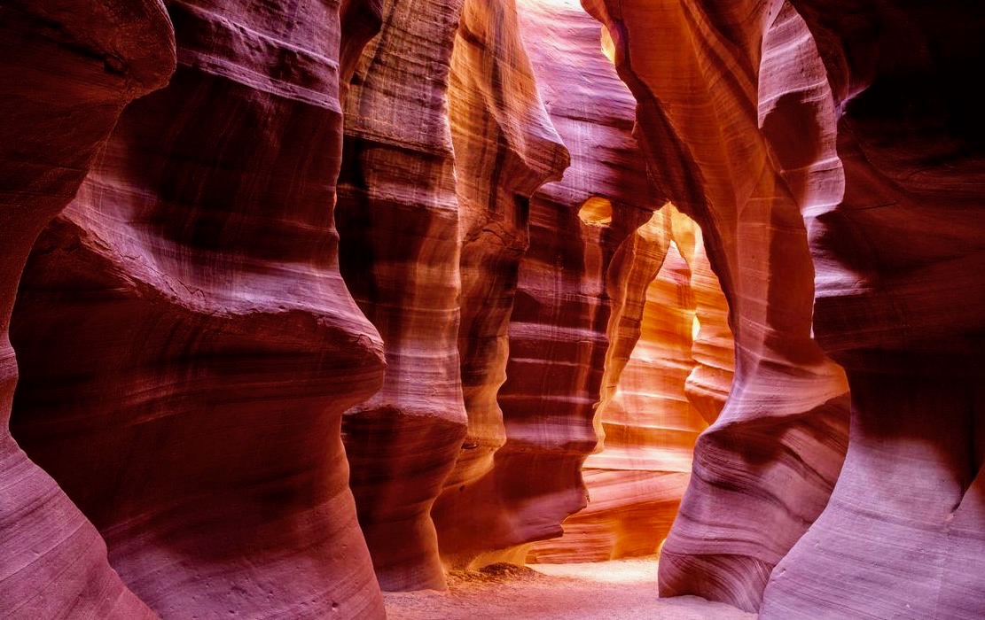 A real photograph of a narrow winding passage in Lower Antelope Canyon with warm orange sandstone walls closing in around a sandy path in Page, Arizona