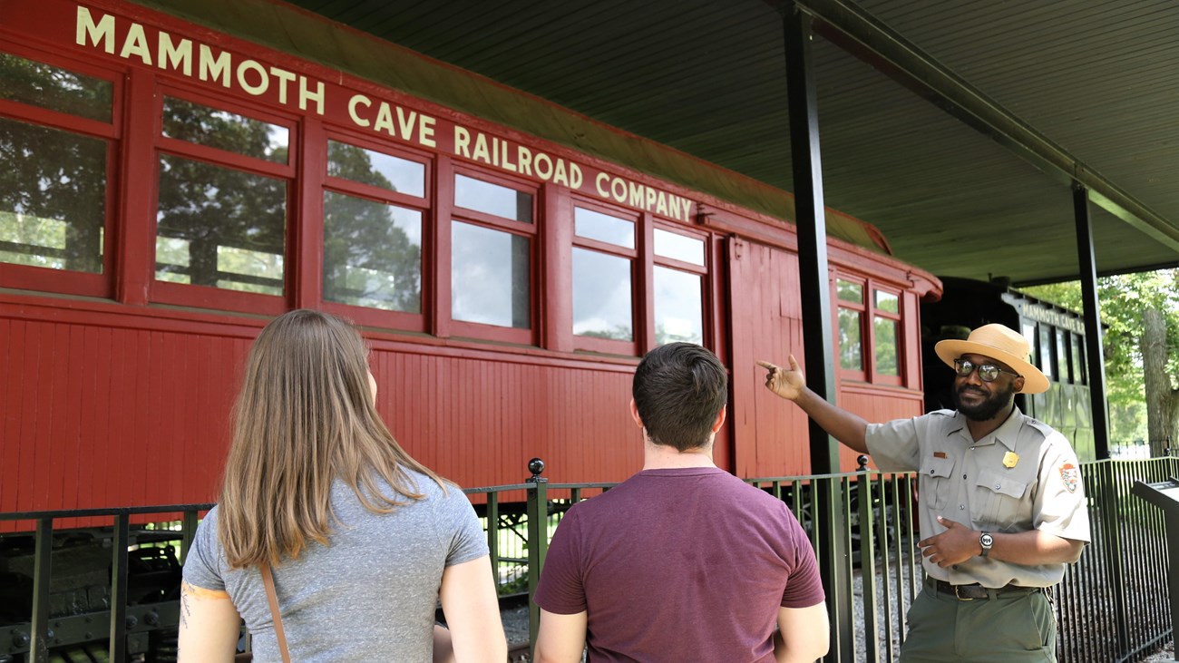 A real photograph of a park ranger leading a small group of visitors along a paved cave path with railings inside Mammoth Cave, warm headlamp and overhead lighting