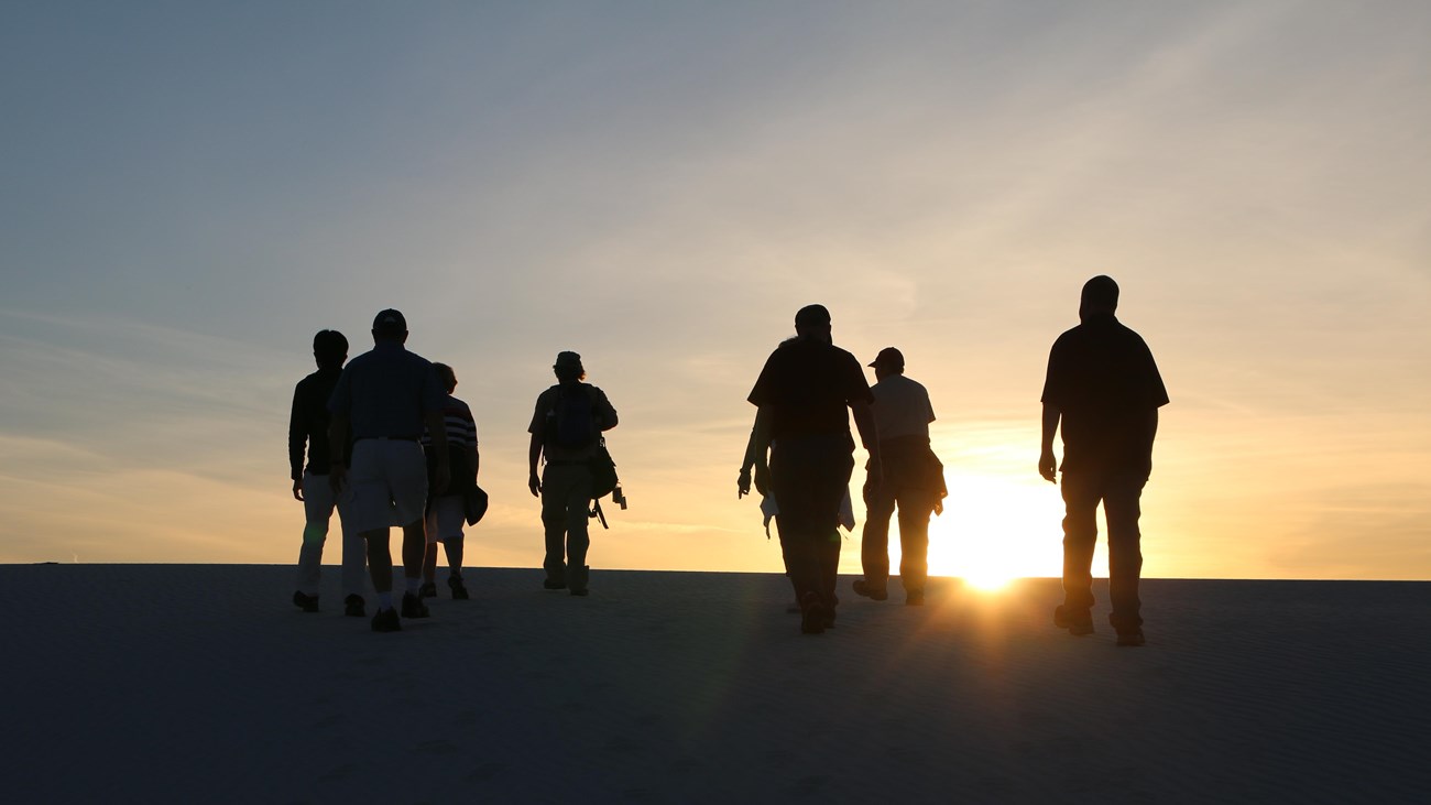 A real photograph of a park ranger speaking to a small group of visitors on white dunes at sunset in White Sands National Park