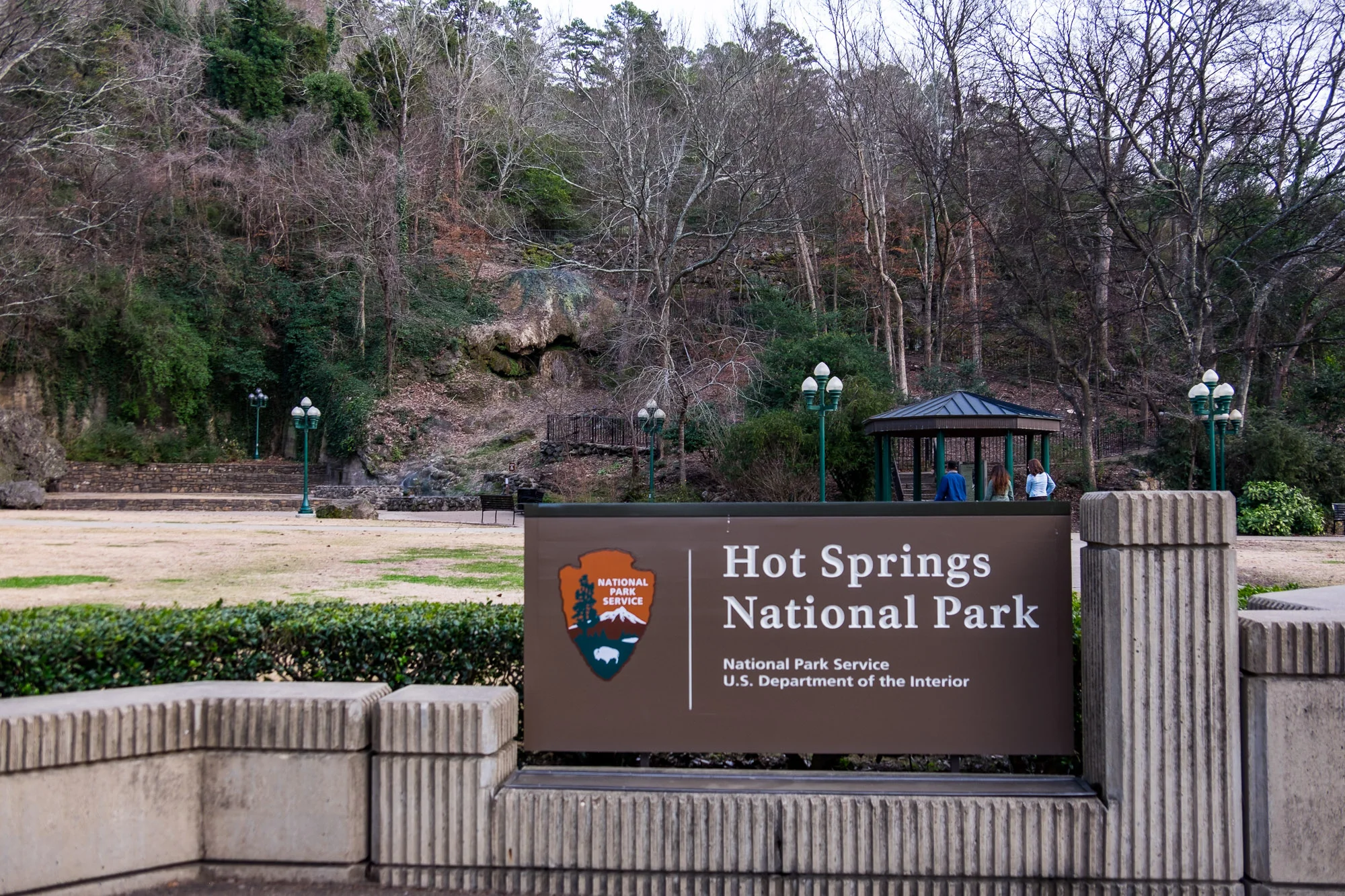 A real photograph of a public thermal spring water spigot in Hot Springs National Park near downtown, with a person holding a reusable bottle and the historic stonework surrounding the fixture