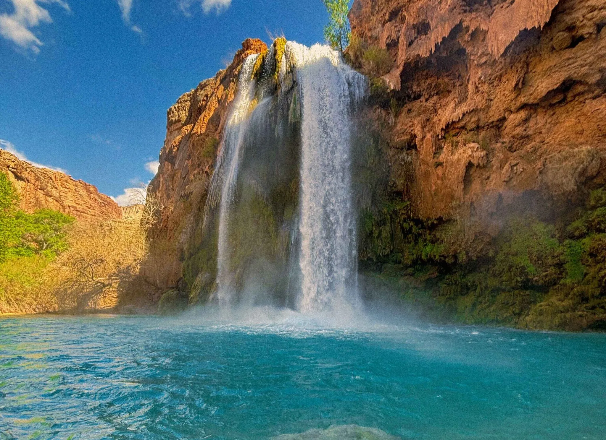 A real photograph of a quiet dirt path through Supai village in the morning light, with simple buildings set back from the road and canyon cliffs rising in the background